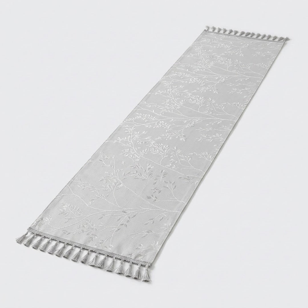 A long, narrow white fabric runner with a subtle tonal floral pattern and tassel fringe on both ends, shown on a light gray background.