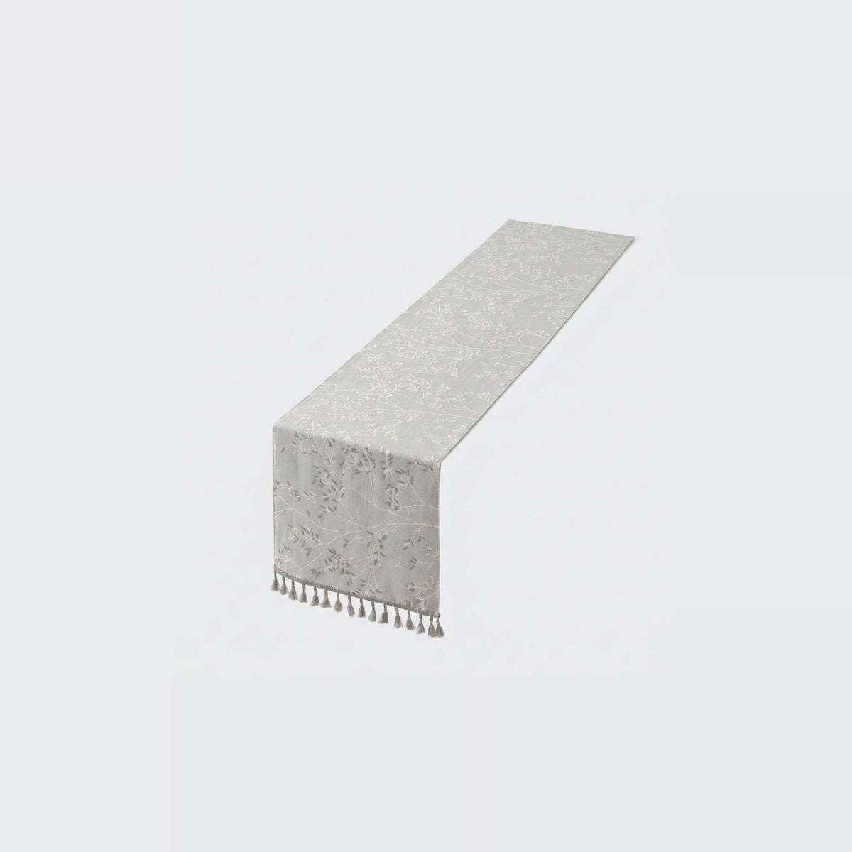 A light gray table runner with a subtle damask pattern and a short fringe of tassels on one end, shown on a plain light gray background.