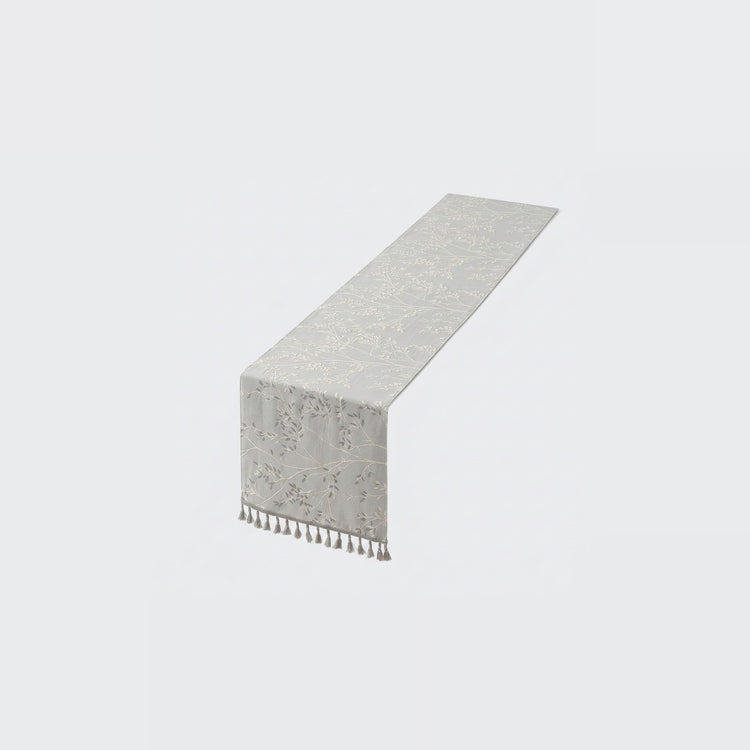 A light gray table runner with a subtle damask pattern and a short fringe of tassels on one end, shown on a plain light gray background.