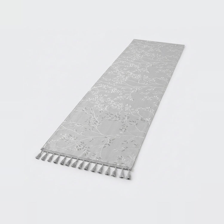 A light gray runner rug with a subtle tonal floral pattern and matching fringe tassels along one end, shown on a clean white background.