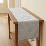 A light gray table runner with an embroidered floral branch pattern and a tassel fringe, draped over a wooden console table in a brightly lit room.