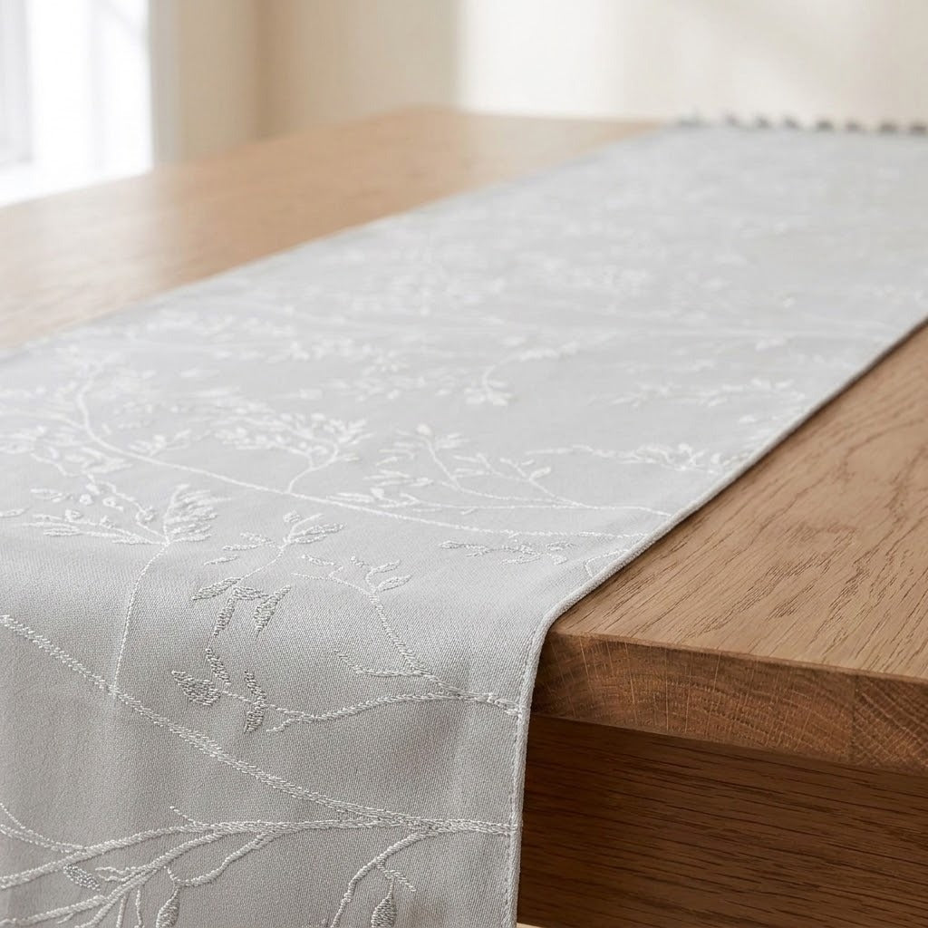 A light gray table runner with delicate white floral embroidery draped over a wooden dining table.