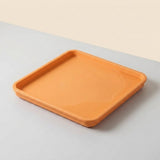 An orange square silicone tray with raised edges, shown on a split beige and gray background.