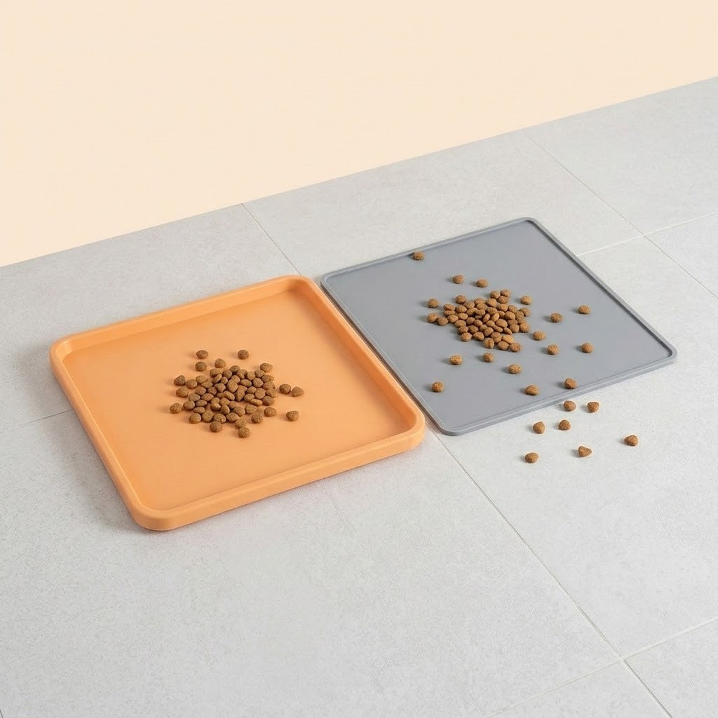 Two square pet feeding mats on a light gray tiled floor, one orange and one gray, each holding a small pile of brown kibble.