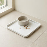A minimalist white rectangular tray with a matching round bowl, placed on a light beige tiled floor near a window. A few brown kibble pieces are scattered on the tray.