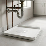 A white rectangular sink tray or drain basin placed on a tiled floor beneath exposed copper and galvanized metal plumbing pipes, with a single water droplet dripping into the center.