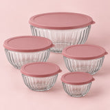 Five clear fluted glass mixing bowls with rose lids centered on a soft pink backdrop.
