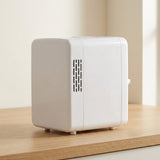 A small white cube-shaped portable heater or cooler with ventilation slots on the side, sitting on a light wooden surface against a soft beige background.