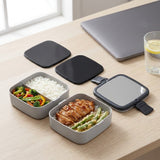 Gray square bento food containers with black lids on a light wood desk, shown with prepared meals and a laptop in a real indoor setting.
