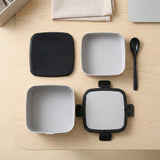 A set of square bento-style containers with lids, shown disassembled on a light wood tabletop. The set includes two light gray containers with black lids, a black lid, and a black spoon, arranged neatly on a beige surface.