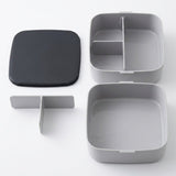 A light gray square bento box shown open with its lid placed to the left. The lid is dark charcoal gray, while the box interior and outer walls are light gray.