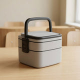 A light gray and black two-tier bento lunch box with a handle, sitting on a light wood table in a softly blurred classroom setting.