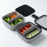 Two gray compartment bento-style lunch boxes with black lids, shown open and filled with broccoli, carrots, cherry tomatoes, and cucumber slices on a clean white background.