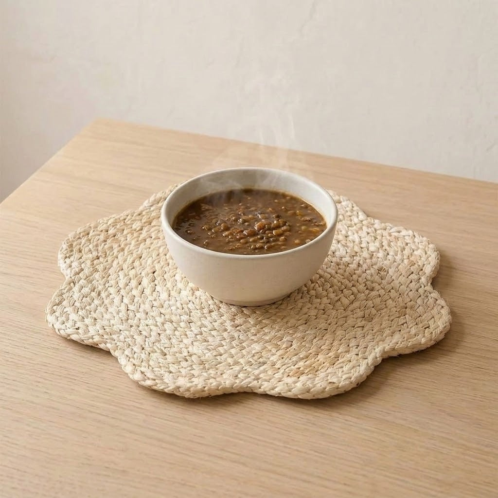 A woven beige scalloped placemat on a light wood table, holding a white ceramic bowl filled with a brown hot soup or stew that has visible steam rising.