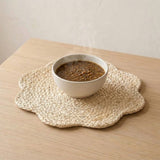 A woven beige scalloped placemat on a light wood table, holding a white ceramic bowl filled with a brown hot soup or stew that has visible steam rising.