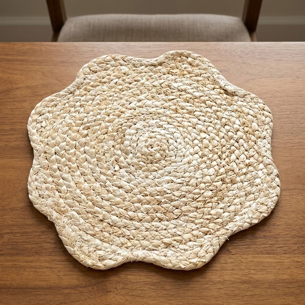 A round braided seagrass placemat with a scalloped edge, displayed on a wooden table with a chair visible in the background.