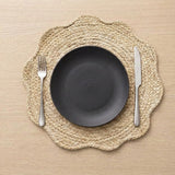 A top-down view of a place setting featuring a scalloped natural jute placemat, a matte black ceramic plate, a silver fork, and a silver knife on a light wood tabletop.
