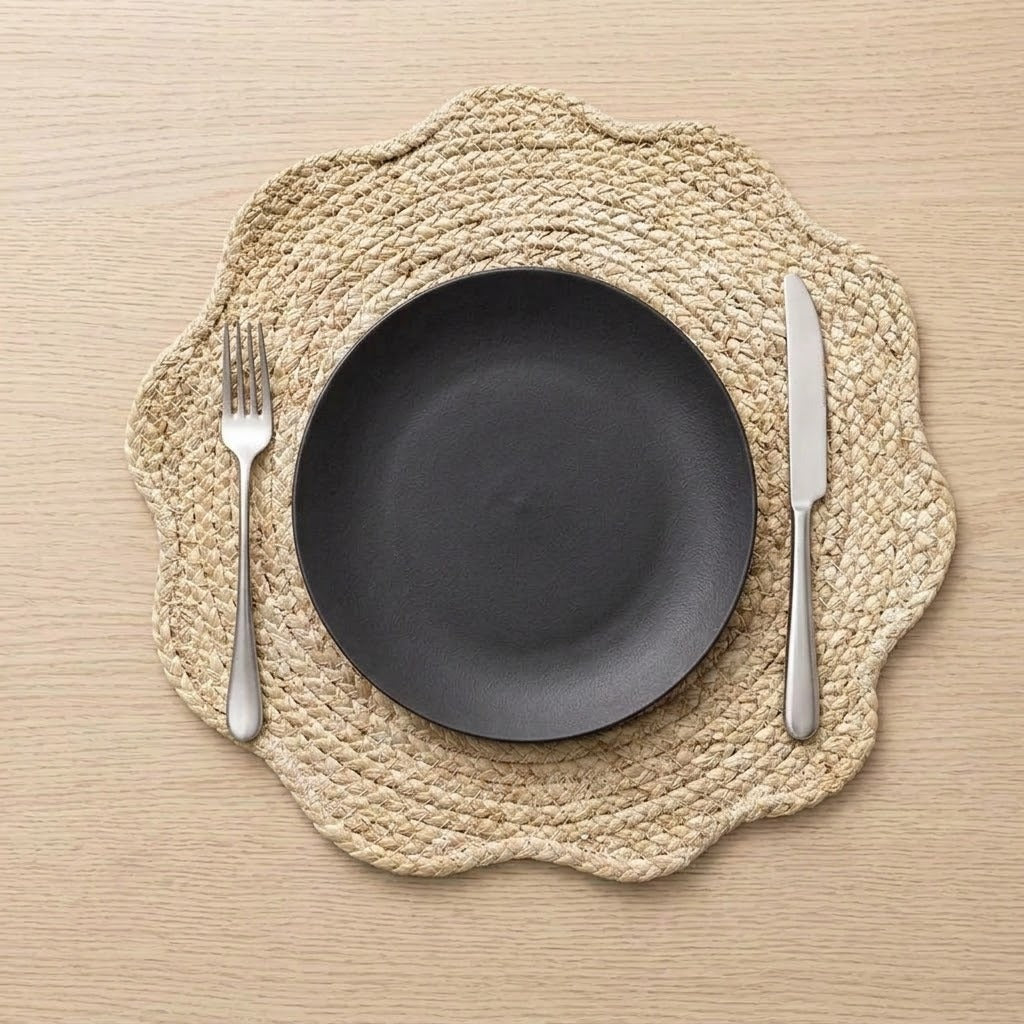 A top-down view of a place setting featuring a scalloped natural jute placemat, a matte black ceramic plate, a silver fork, and a silver knife on a light wood tabletop.