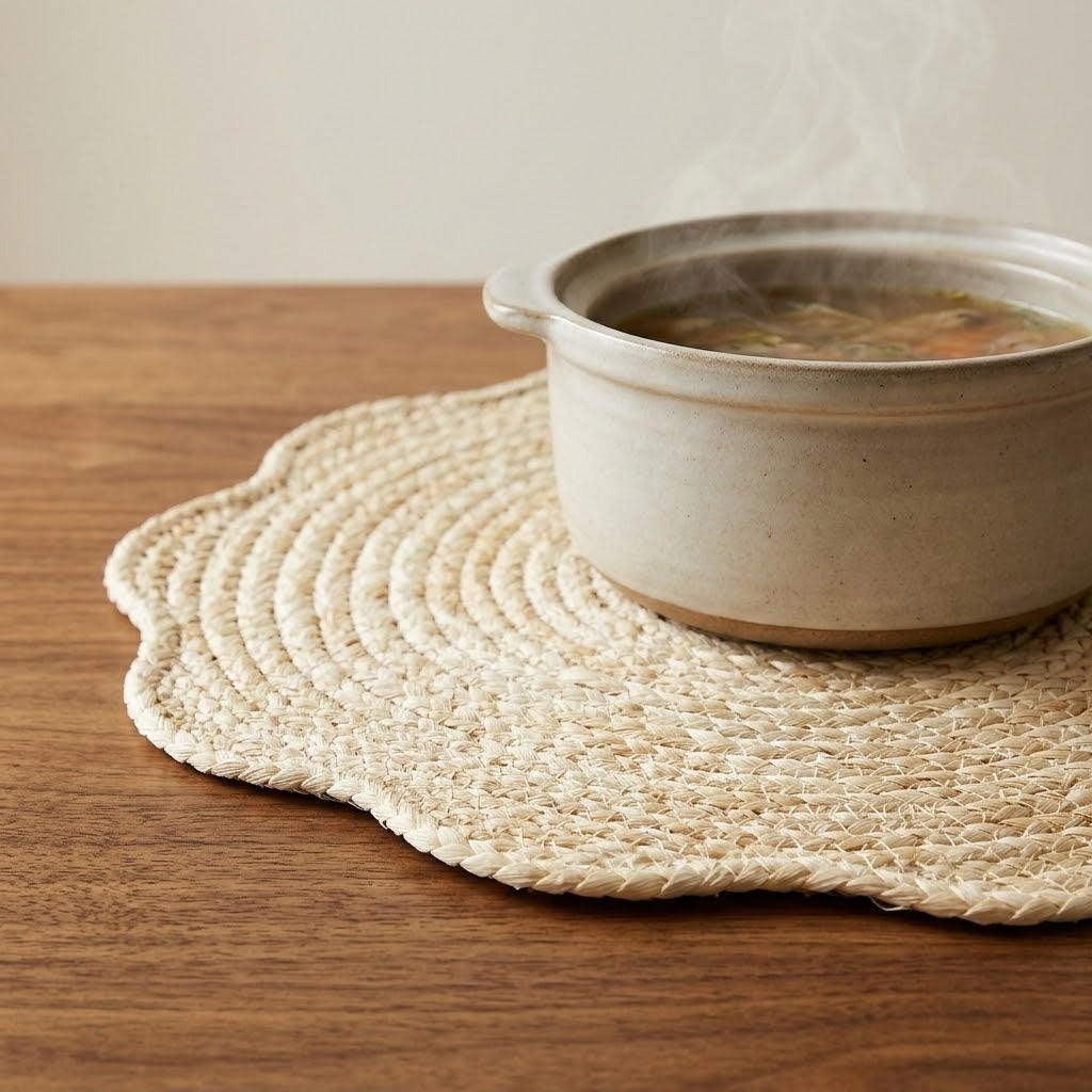 A woven round placemat with scalloped edges sits on a wooden table, partially covered by a beige ceramic pot of soup with visible steam rising.