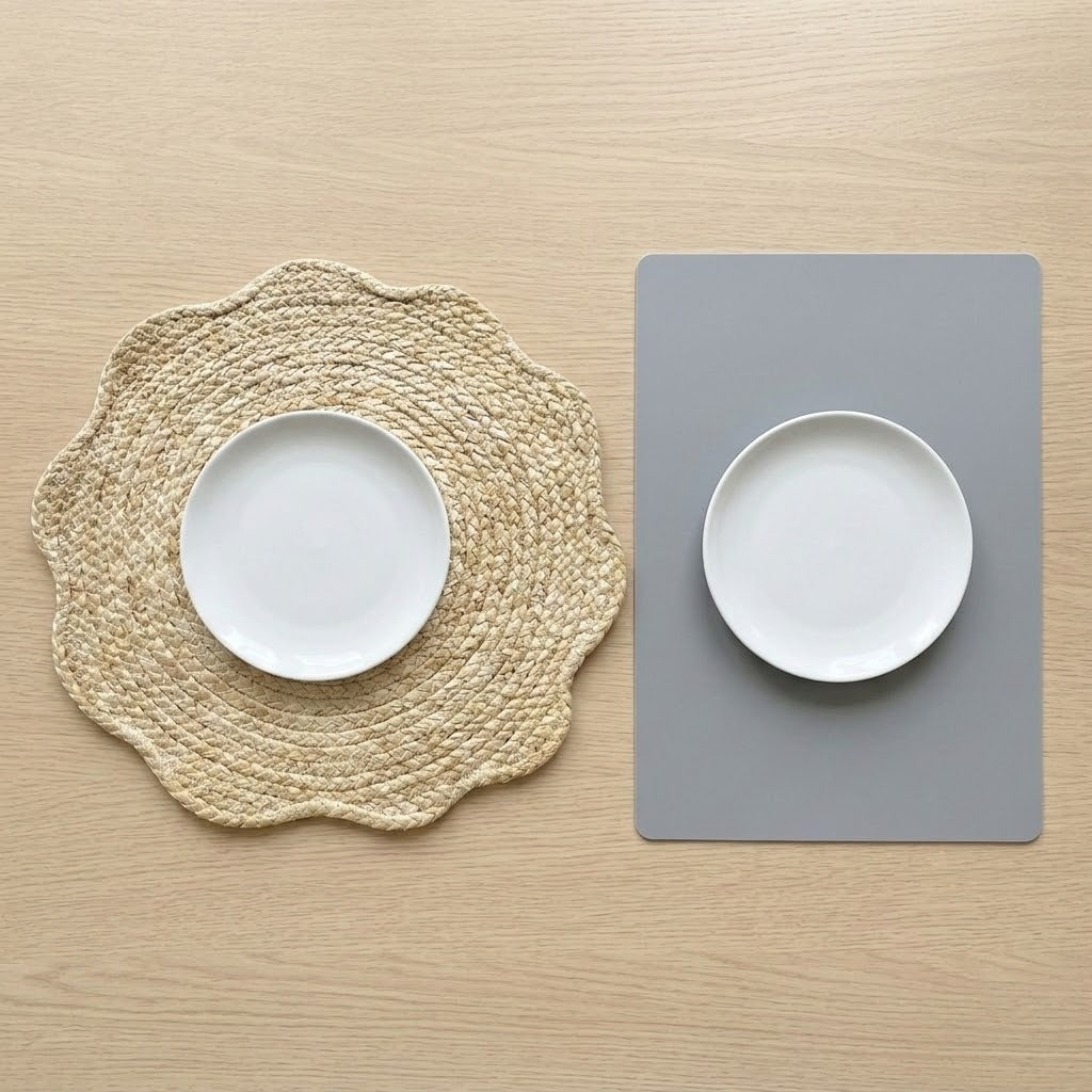 Two place settings on a light wood tabletop: a white oval plate on a round woven beige placemat on the left, and a white oval plate on a rectangular gray placemat on the right.