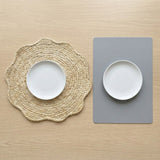Two place settings on a light wood tabletop: a white oval plate on a round woven beige placemat on the left, and a white oval plate on a rectangular gray placemat on the right.