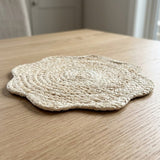 A round woven straw placemat with a scalloped edge sits on a light wood table in a softly blurred indoor setting.