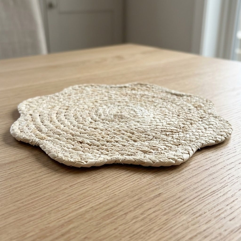 A round woven straw placemat with a scalloped edge sits on a light wood table in a softly blurred indoor setting.