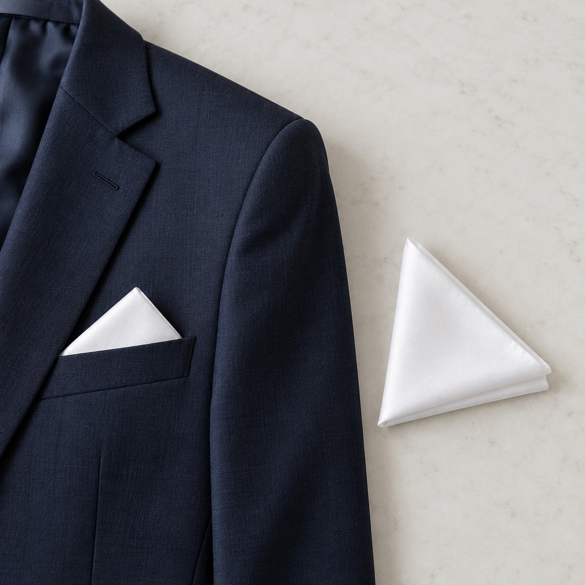 A folded white polyester pocket square rests beside a suit jacket pocket to show its compact scale.