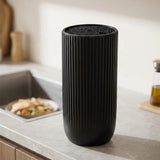 A tall black ribbed cylindrical container or vase with a textured top, standing on a light gray kitchen countertop near a sink.