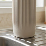 A ribbed off-white cylindrical container or dispenser placed on a stainless steel sink counter in a kitchen setting.
