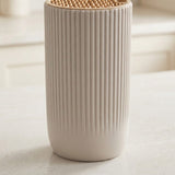 A ribbed ceramic toothbrush holder with a natural wood lid and visible bristles, standing on a light countertop in a softly blurred kitchen setting.