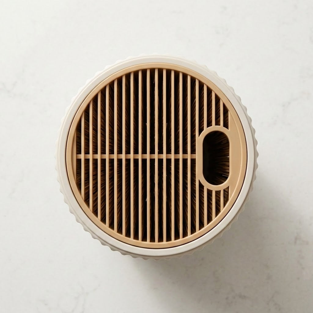 Top-down view of a round pet grooming brush or vacuum cleaner head with a beige and white textured outer body, a beige slatted face, and a dark brown central opening, shown on a light marble surface.
