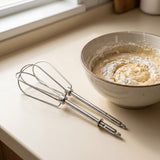 A stainless steel hand mixer whisk resting on a beige kitchen countertop next to a ceramic bowl containing a pale batter or cream with a dusting of flour.