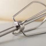 Close-up of a stainless steel electric hand mixer attachment showing the metal beaters and central hub, set against a soft beige and white background.