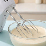 A white electric hand mixer with two stainless steel beaters is mixing a pale creamy liquid in a clear glass bowl, set against a softly blurred kitchen background.