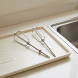 Two stainless steel electric mixer beaters resting on a beige plastic sink tray next to a stainless steel sink.