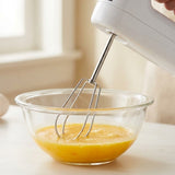 A white electric hand mixer with metal wire beaters is being used to mix a yellow-orange liquid in a clear glass bowl on a light countertop near a window.
