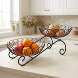 A black metal two-tier fruit and bread stand on a kitchen counter, with apples and oranges in the lower bowl and bread in the upper bowl. The background shows a softly blurred kitchen interior with white cabinets and a window.