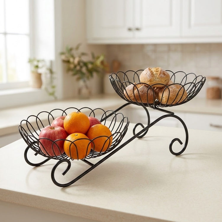 A black metal two-tier fruit and bread stand on a kitchen counter, with apples and oranges in the lower bowl and bread in the upper bowl. The background shows a softly blurred kitchen interior with white cabinets and a window.