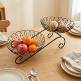 A black metal two-tier fruit and bread basket displayed on a wooden dining table, holding red apples, oranges, and pastries. The scene is styled with plates, napkins, and silverware in a warm, domestic setting.