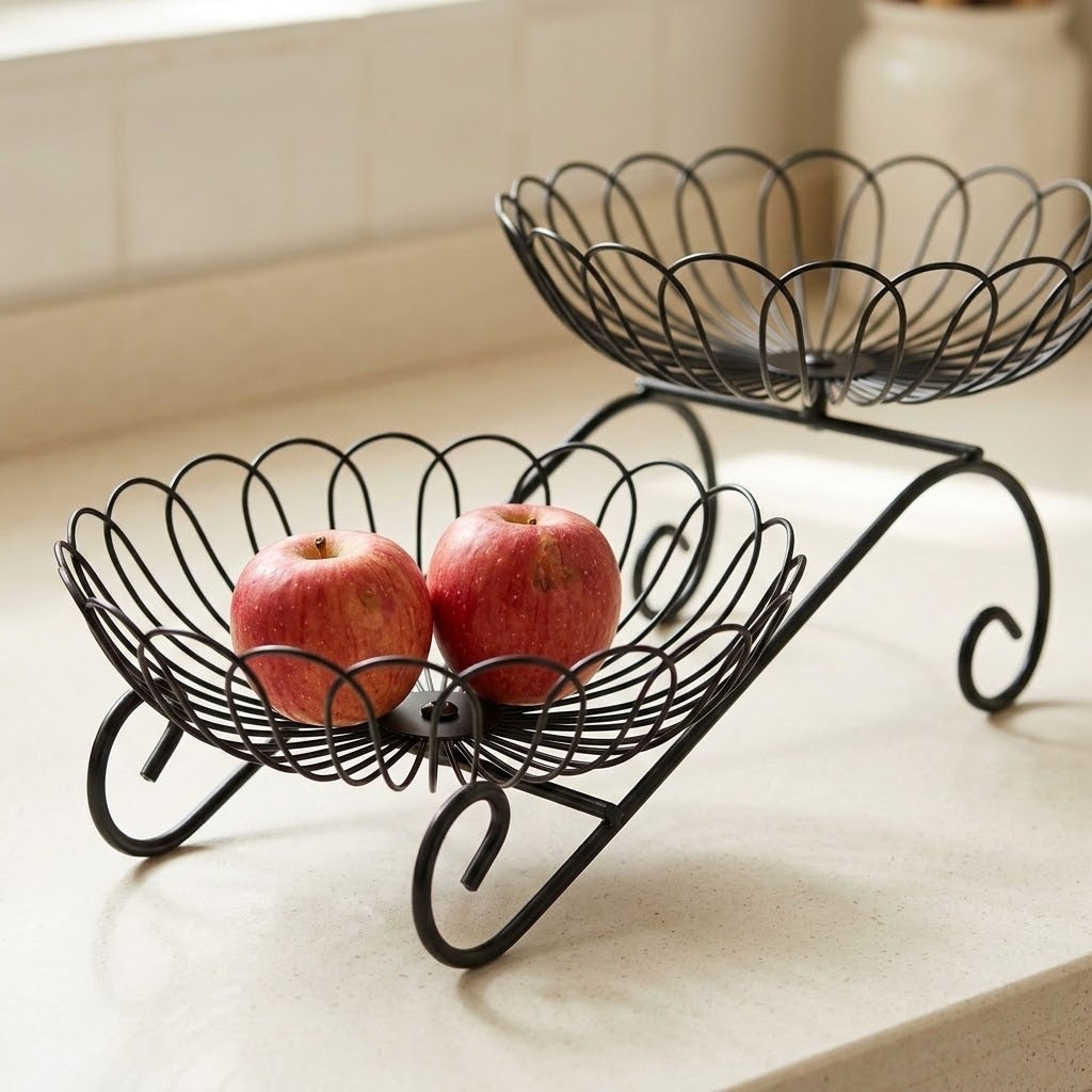 A black metal two-tier fruit basket with decorative scalloped wire bowls, shown on a light countertop with two red apples in the lower bowl.