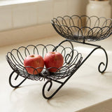 A black metal two-tier fruit basket with decorative scalloped wire bowls, shown on a light countertop with two red apples in the lower bowl.