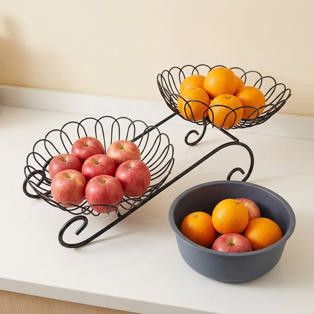 A two-tier black metal wire fruit basket holding red apples and oranges, shown on a white countertop next to a dark gray bowl with fruit, against a beige wall.