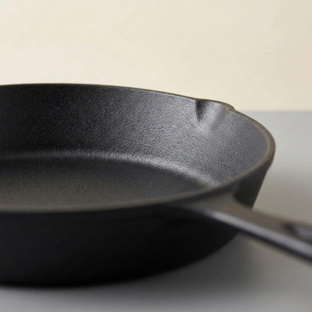 A close-up of a black cast iron skillet showing its textured interior and a portion of the handle, set against a neutral light gray and beige background.
