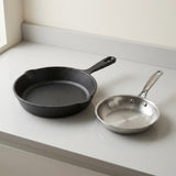 A black cast iron skillet and a small stainless steel saucepan resting on a light gray countertop in a clean kitchen setting.