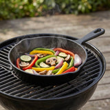 A black cast iron skillet filled with colorful sliced vegetables, including zucchini, bell peppers, onions, and mushrooms, sitting on a black kettle-style grill outdoors.