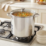 A large stainless steel stockpot with a glass lid sits on a gas stovetop in a bright kitchen. The pot is filled with a colorful vegetable stew, and two cream-colored bowls are stacked on the counter to the right.