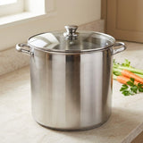 A large stainless steel stockpot with a glass lid and side handles sits on a light stone countertop in a kitchen setting, with carrots and parsley visible to the right.