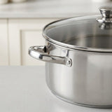 A stainless steel stockpot with a matching glass lid and chrome handle, shown on a light gray countertop with a blurred kitchen background.