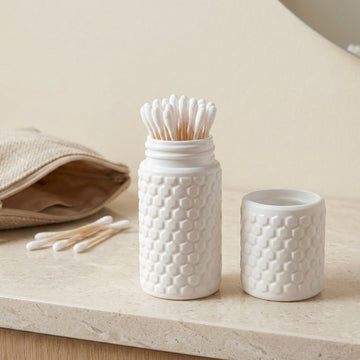 A white textured cotton swab jar with wooden cotton swabs inside and its matching lid beside it, set on a light stone surface with a beige fabric pouch in the background.